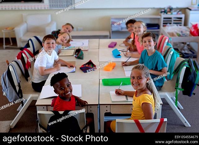 High angle view of a diverse group of schoolchildren sitting at desks working in an elementary school classroom, turning around and smiling to camera
