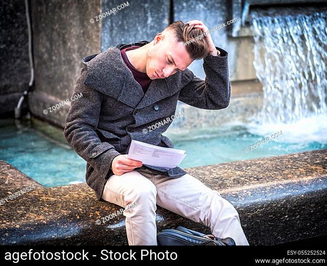Young man, a student, sitting by fountain reading paper sheets in city setting in a winter day