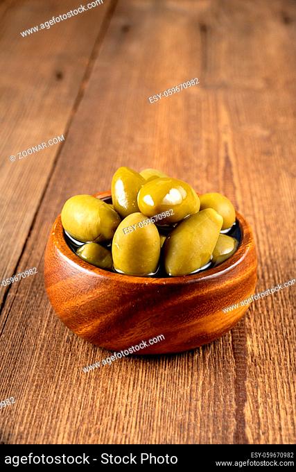 Green olives in olive oil in a small wooden bowl on a rustic wooden background
