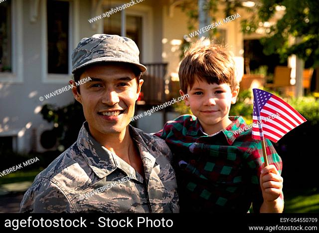 Portrait of a young adult mixed race male soldier in the garden outside his home, carrying his son, who is holding a US flag, both smiling to camera
