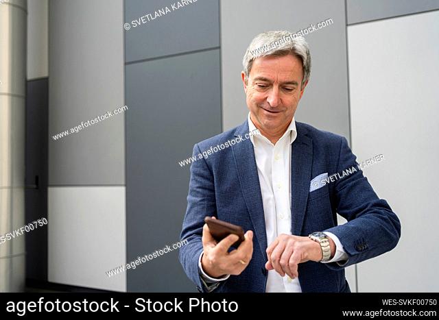 Mature businessman holding mobile phone and checking time on wristwatch in front of gray wall