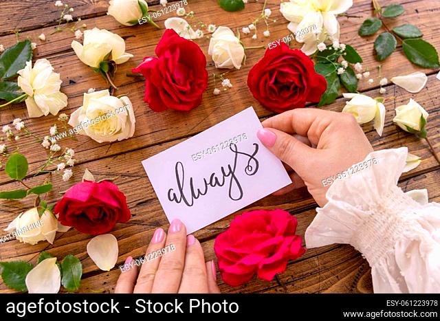 Hands with handwritten card ALWAYS surrounded by red and cream roses close up on a wooden table. Femminine romantic declaration of love near flowers