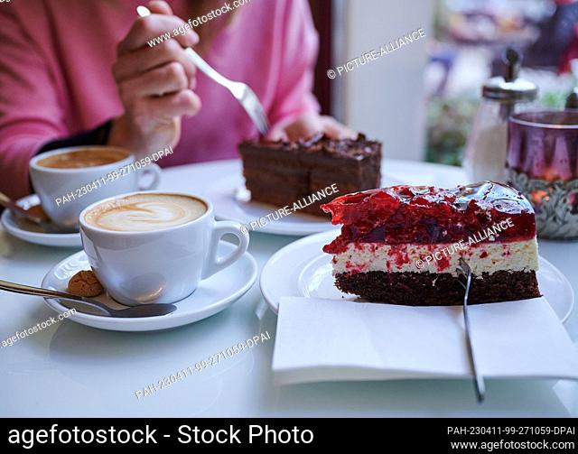 PRODUCTION - 27 March 2023, Berlin: A woman stabs a piece of cake with a cake fork while another piece of cake and two cappuccini sit on the table at a café in...