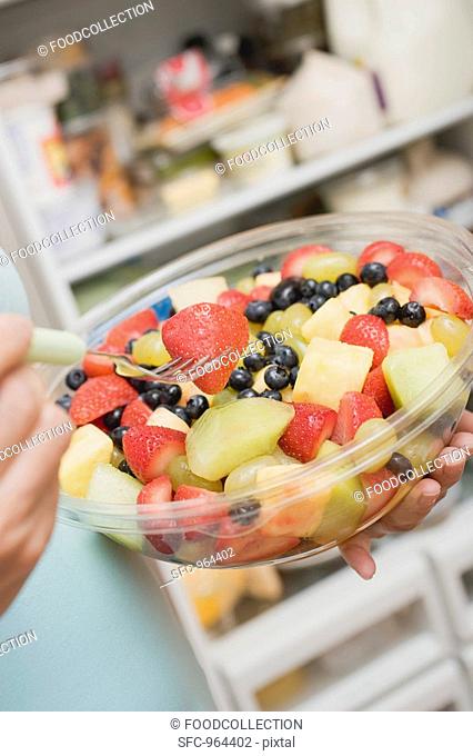 Woman eating fruit salad out of plastic bowl in front of refrigerator