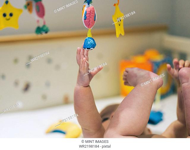 A baby girl in a diaper lying in a cot reaching up to a colourful mobile hanging above her