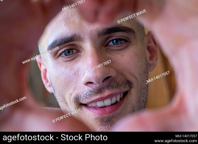 cheerful affectionate teenager man take romantic selfie portrait looking at camera through heart of joined fingers. enamored young boy showing his love to the...