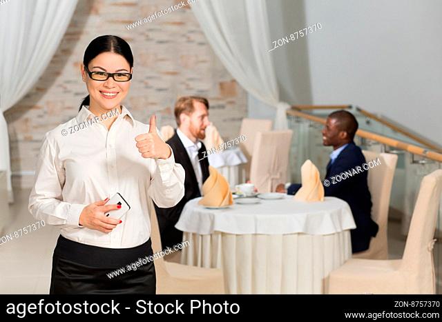 Happy business woman showing thumb-up in restaurant while her colleagues or partners communicating on background at table