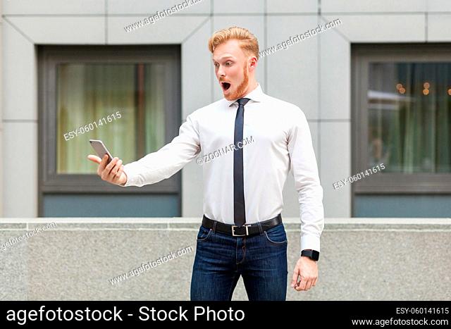 Ginger young adult business person looking at screen smart phone with shocked face. Outdoor shot