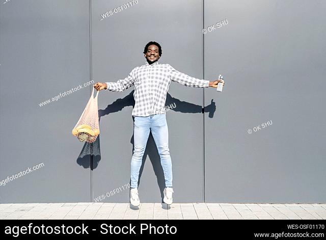 Cheerful jumping in front of gray wall holding mesh bag