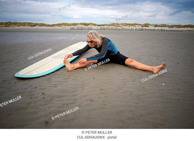 Senior woman sitting on sand, stretching, surfboard beside her