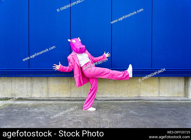 Woman wearing vibrant pink suit and hippo mask posing on one leg in front of blue wall