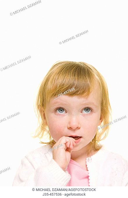 A three year old girl with blonde hair wearing a white sweater and a pink top looks upwards with a concerned expression while biting on her fingers