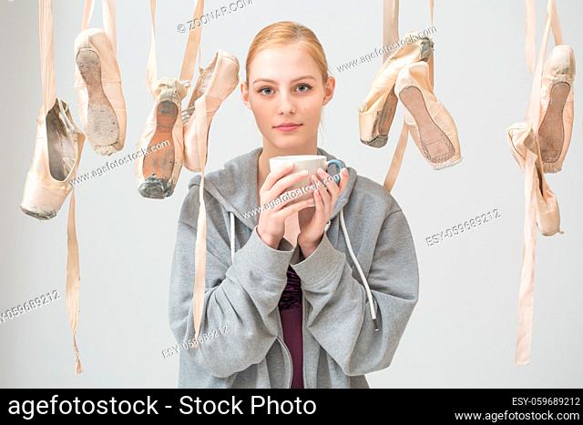 Portrait of a young ballerina with a cup of tea in her hands is among the hanging ballet slippers. Pointe shoes are arranged in random order