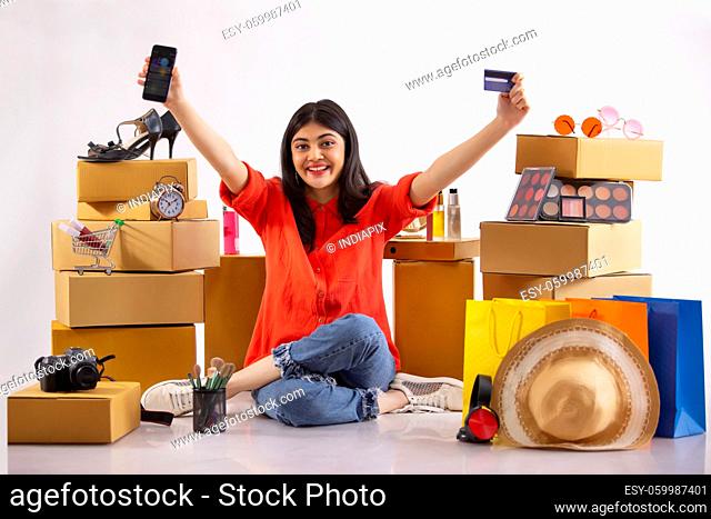 A young woman sitting with smartphone and credit card amidst shopping items in the background