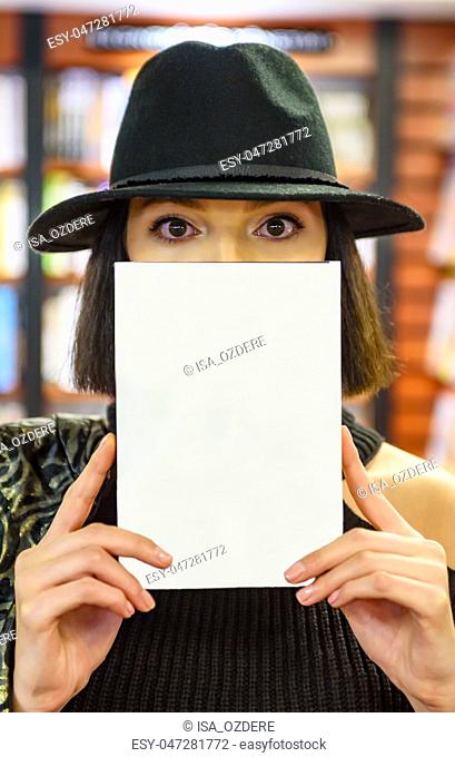 Attractive beautiful woman with black hat holding book partially covered face with closed book and looking at camera