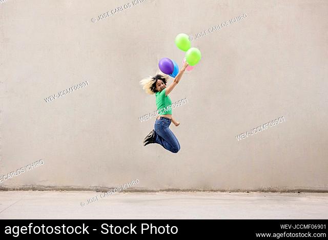 Happy young woman with multi colored balloons jumping in front of wall