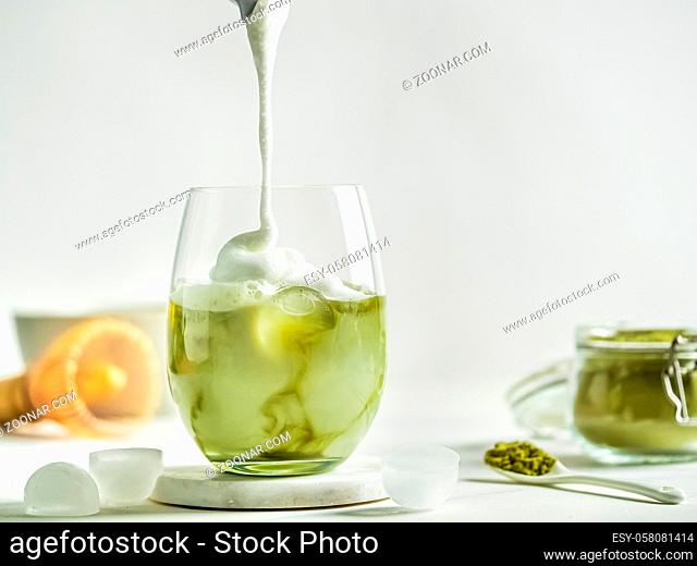 Iced Matcha Latte Tea with pouring whipped cream in tumbler glass. Matcha latte and ingredients on white marble background