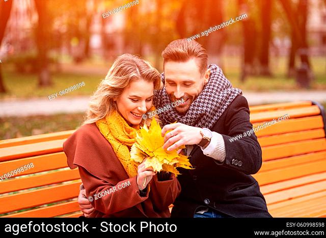 Young embracing happy romantic couple sitting gently hugged on a bench in park wearing coats and scarfs. A romantic couple sitting on a bench hugging in the...