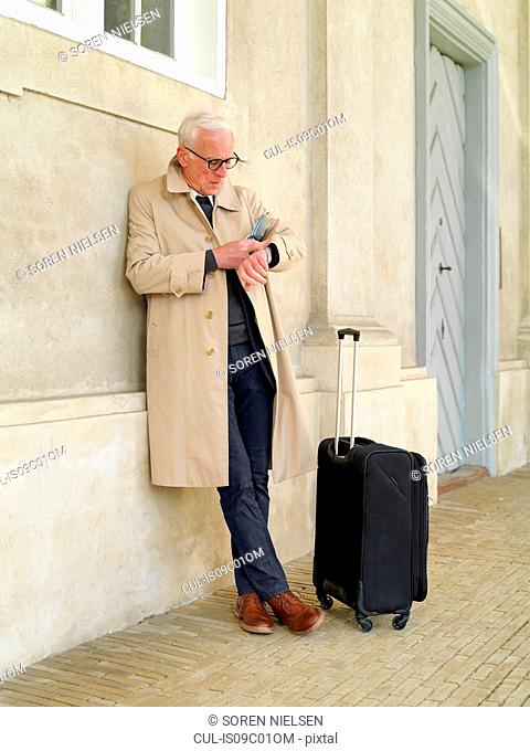 Senior man with wheeled suitcase leaning against building looking at his watch, Copenhagen, Hovedstaden, Denmark