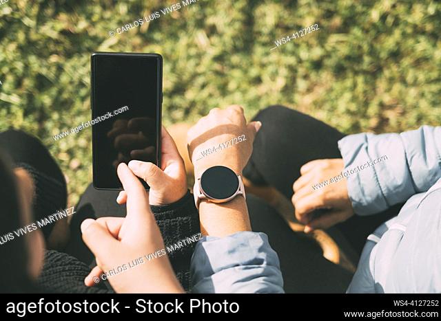 Hispanic male teenager holding smartphone looking away while sitting on grass with Hispanic mother and sister in park on sunny day,