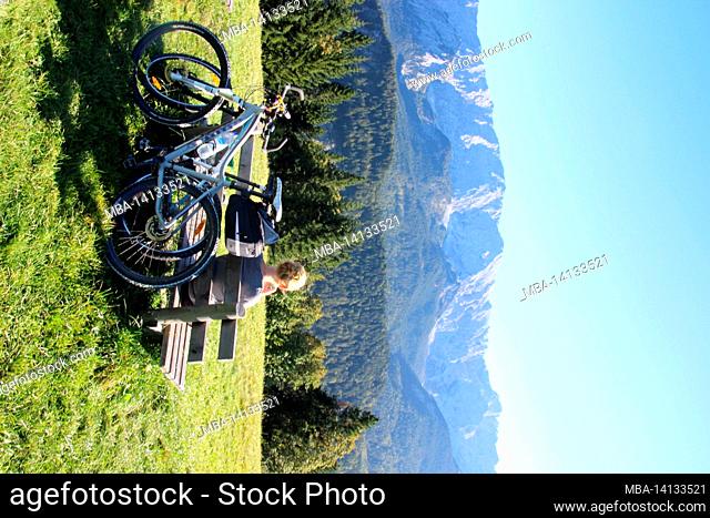 young woman takes a break on a bicycle tour on a bench at the eckbauer in front of the wetterstein massif with a wonderful blue sky, autumn in werdenfelser land