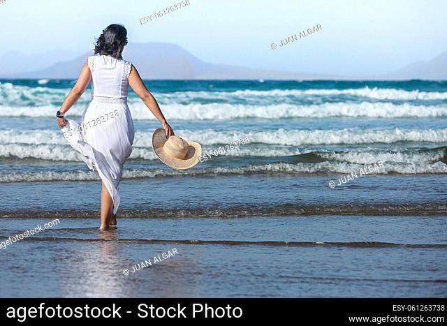 Back view of anonymous woman in white dress carrying straw hat and walking in waves of stormy sea against blue sky while spending summer vacation on beach