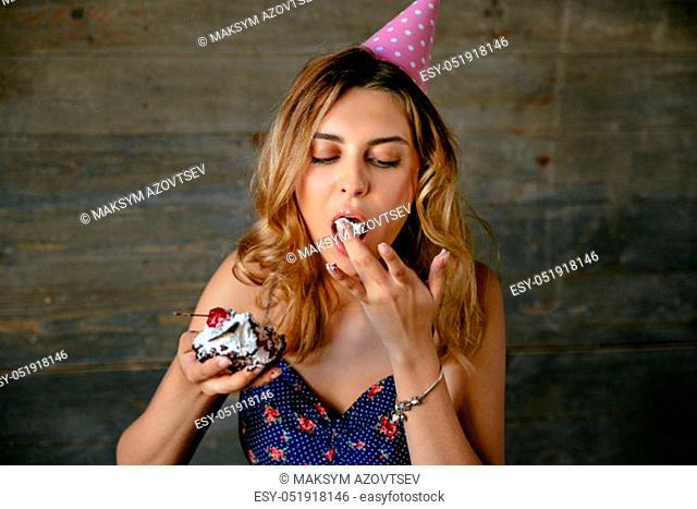 Gorgeous young woman eating cream of chocolate cake from her fingers, celebrating her birthday, wearing holiday hat