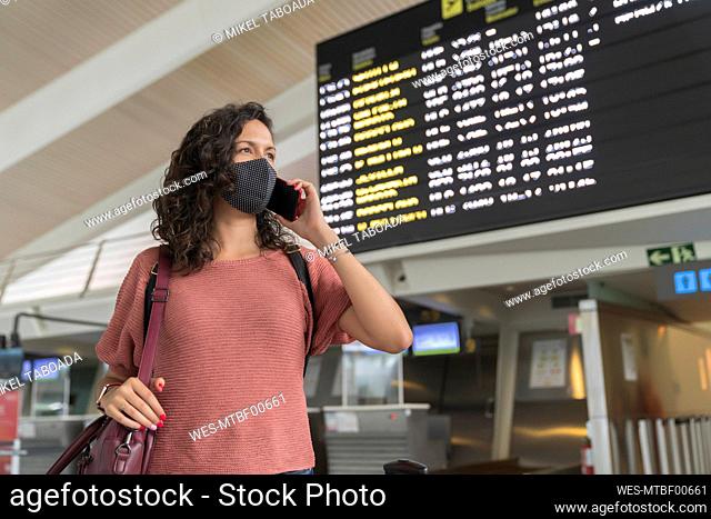 Young woman with protective face mask talking on smart phone while standing at airport