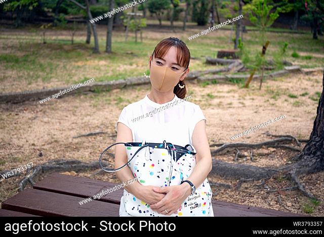 A mature Japanese woman outdoors in a park, wearing a face mask, holding a bag