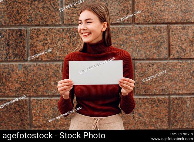 Young woman smiling with smile holds white paper in hand stone background. Girl with white blank template sheet with empty space
