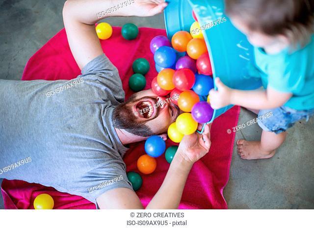 Father lying on floor, toddler pouring bucket of balls over him