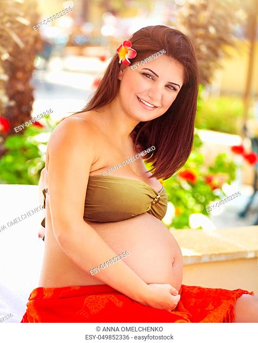 Portrait of a beautiful pregnant woman sitting on the beach, future mother spending summer holidays on a tropical resort, happy healthy pregnancy