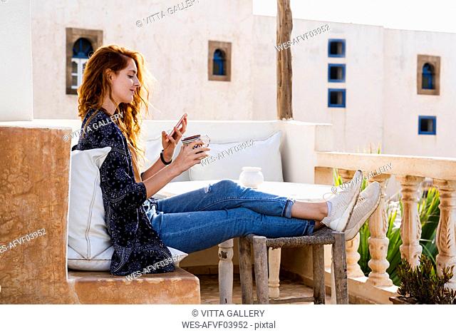 Smiling redheaded young woman relaxing with cup of coffee on roof terrace looking at cell phone