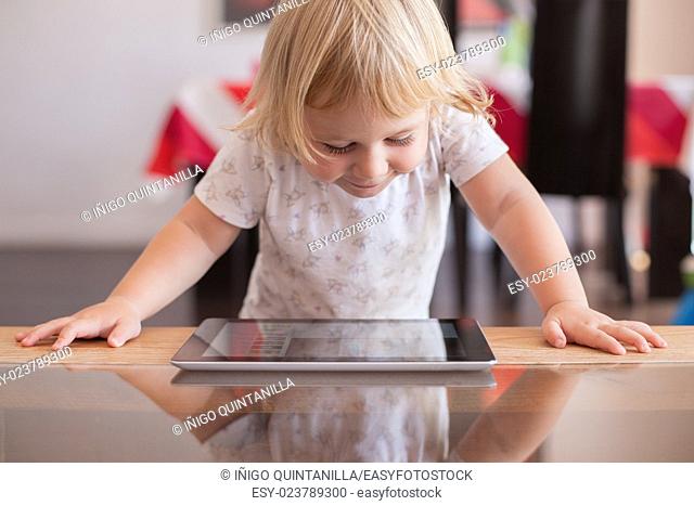 blonde two years old baby white shirt smiling happy face reading and watching digital tablet on wood and crystal table indoor at home