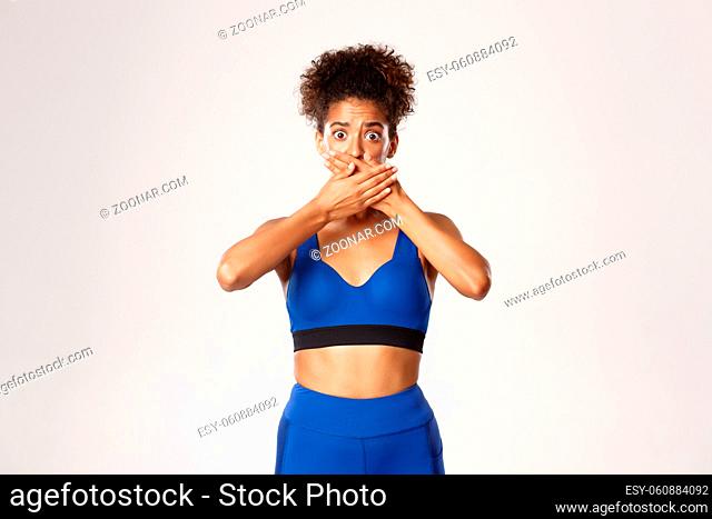 Waist-up of scared and shocked african-american female athlete, shut her mouth with hands, holding scream, standing against white background