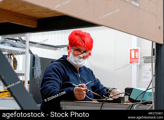 Industrial worker wearing a face mask due to a coronavirus pandemic solders cables from factory production equipment. High quality photo