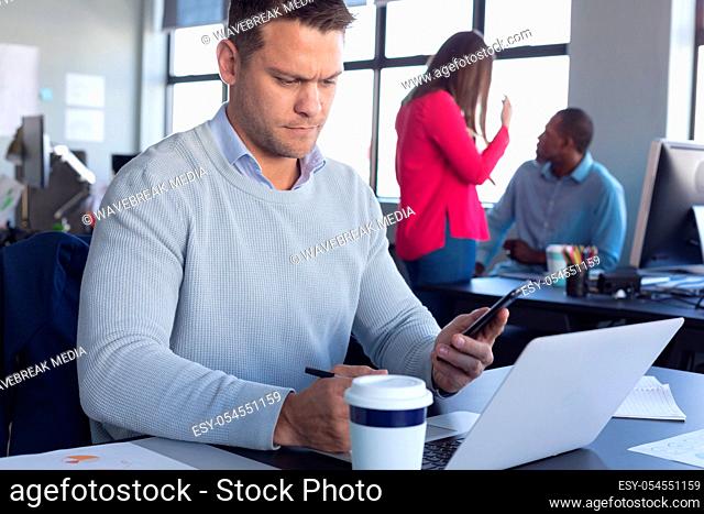 Front view close up of a Caucasian male business creative working in a casual modern office, using a laptop and looking at his smartphone