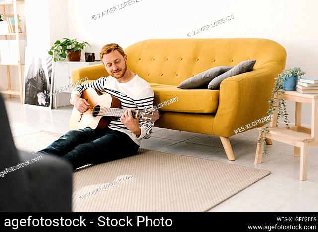 Young man with red hair practicing guitar sitting in front of sofa at home