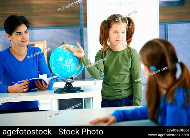 Young schoolgirl pointing at globe in class, looking at another pupil, teacher smiling