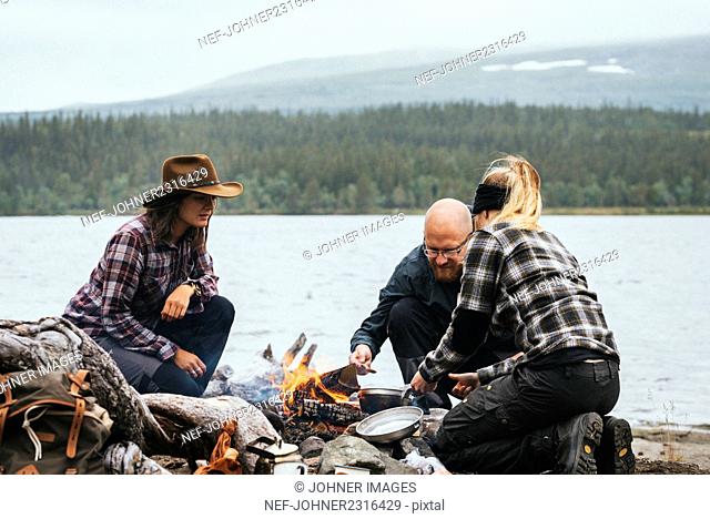 Three people cook food by fire