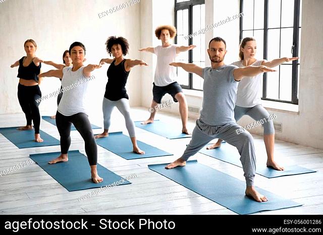 Young diverse people wearing sportswear doing Warrior two exercise at group lesson, practicing yoga, standing in Virabhadrasan pose on mats