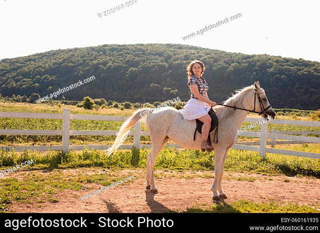 The young beautiful woman in a white skirt is sitting on a white horse. She holds the horse by the rein