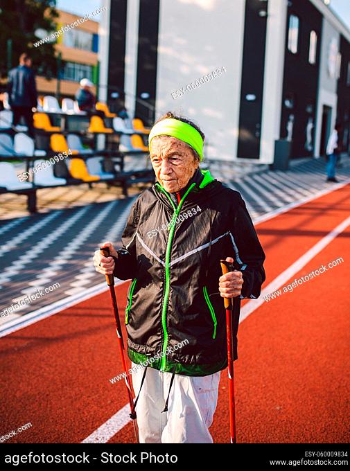 Old woman in sportswear practicing nordic walking outdoors on rubber treadmill in stadium. Older female walk by scandinavian walk use trekking sticks and nordic...