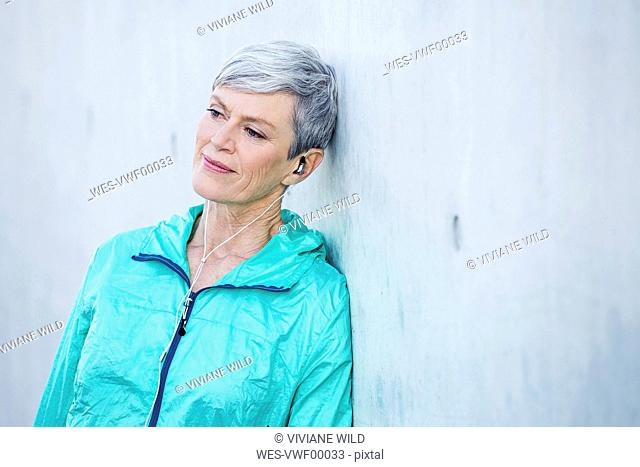 Smiling sporty mature woman with earbuds leaning against concrete wall