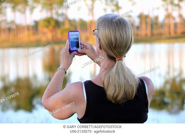 A senior woman taking scenic pictures beside a lake with a camera phone, ipod or iphone