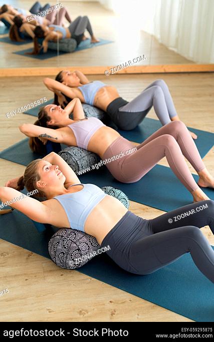 Restorative yoga with a bolster. Group of three young sporty attractive women in yoga studio, lying on bolster cushion, stretching and relaxing during...