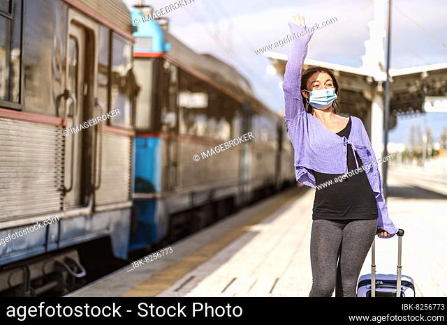 A young woman in mask and with luggage greetings somebody at the platform of train station
