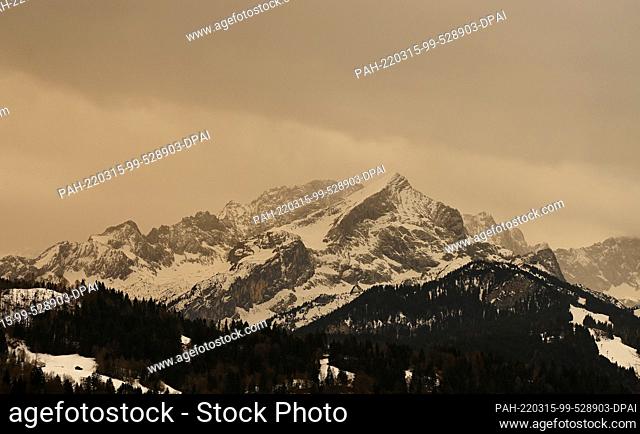 15 March 2022, Bavaria, Krün: Over the peaks of the Wetterstein mountains bad weather with Sahara dust is coming up that colors the sky yellow/orange