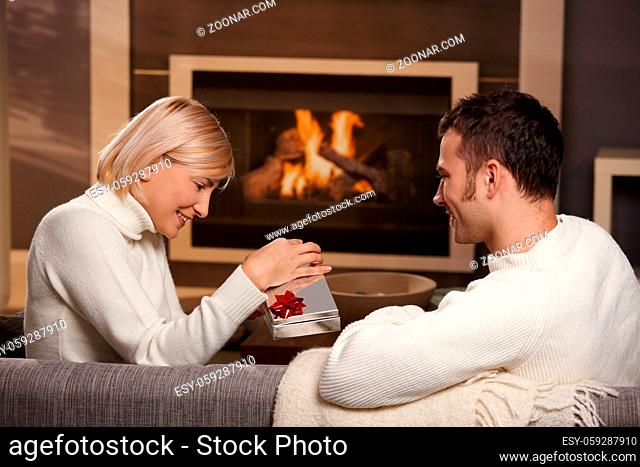 Young romantic couple sitting on couch in front of fireplace at home, man giving gift, side view