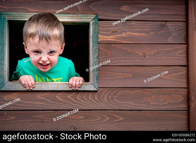 Portrait of a cute cheeky little Caucasian boy looking through the window of a wooden toy house in a outdoor playground